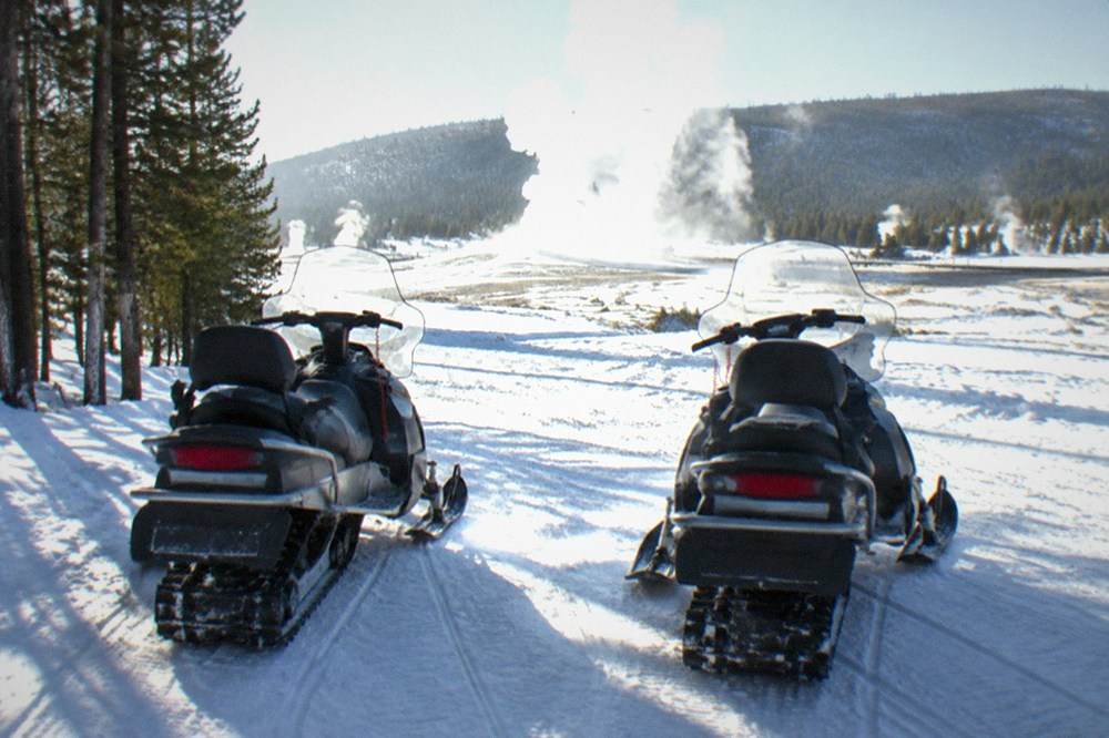 Two touring snowmobiles parked side by side on a wide groomed snow road with steaming thermal pools in the distance