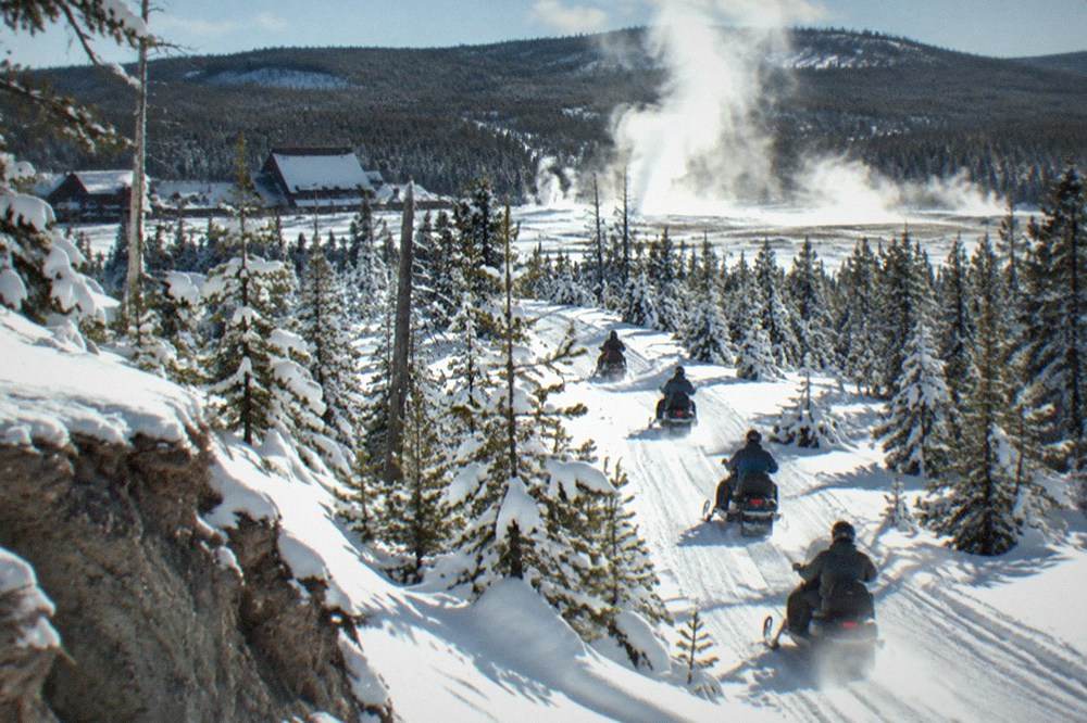 A line of snowmobiles traveling a groomed snow road through snowy pine forest toward a steaming geyser basin