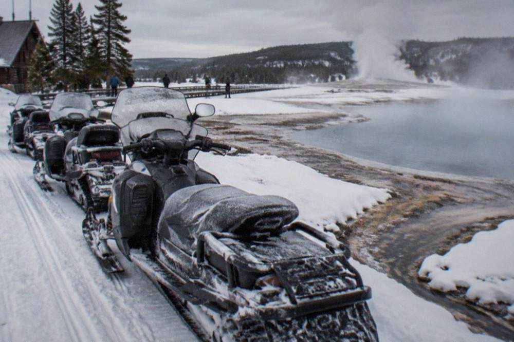 A line of snowmobiles parked on a groomed snowy road beside steaming thermal pools in a winter geyser basin
