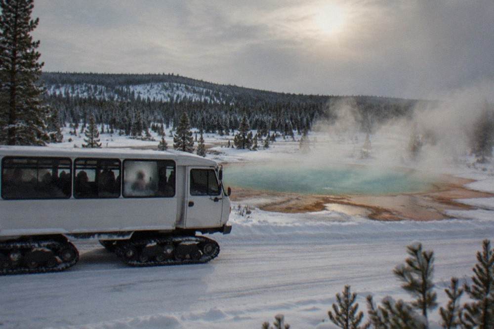 A snowcoach passes a steaming geothermal pool on a winter road in Yellowstone