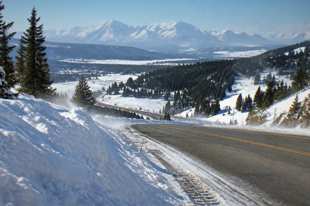 Plowed winter highway winding between snowbanks toward distant mountain peaks