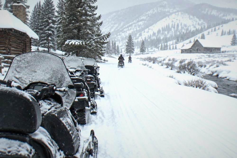 A line of parked snowmobiles on a groomed white road winding between snowy hills