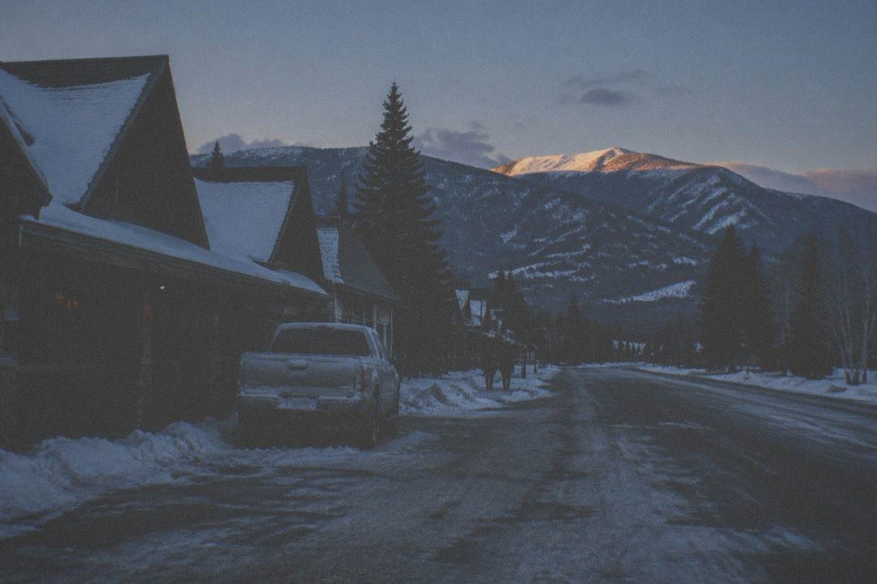 Snow-covered gateway town street with mountains rising behind the rooftops at dusk