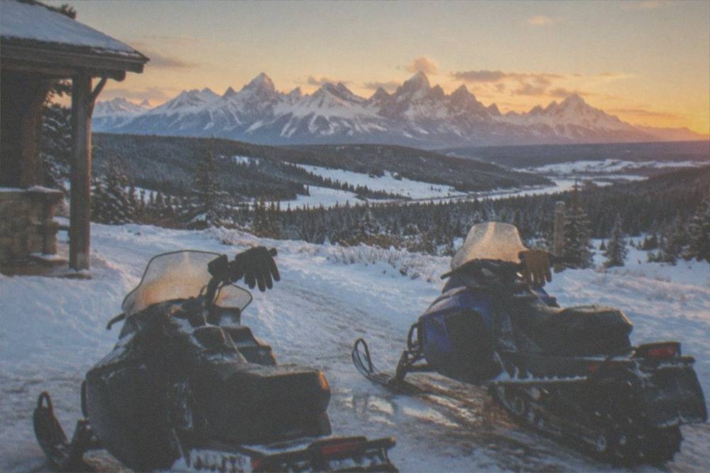 Two snowmobiles parked beside a snowy trail with mountains in the distance