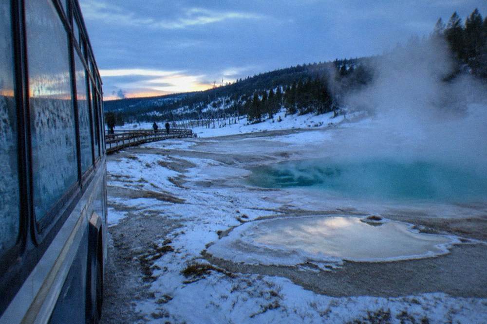 a snowcoach with large windows parked beside steaming thermal pools in a snowy valley