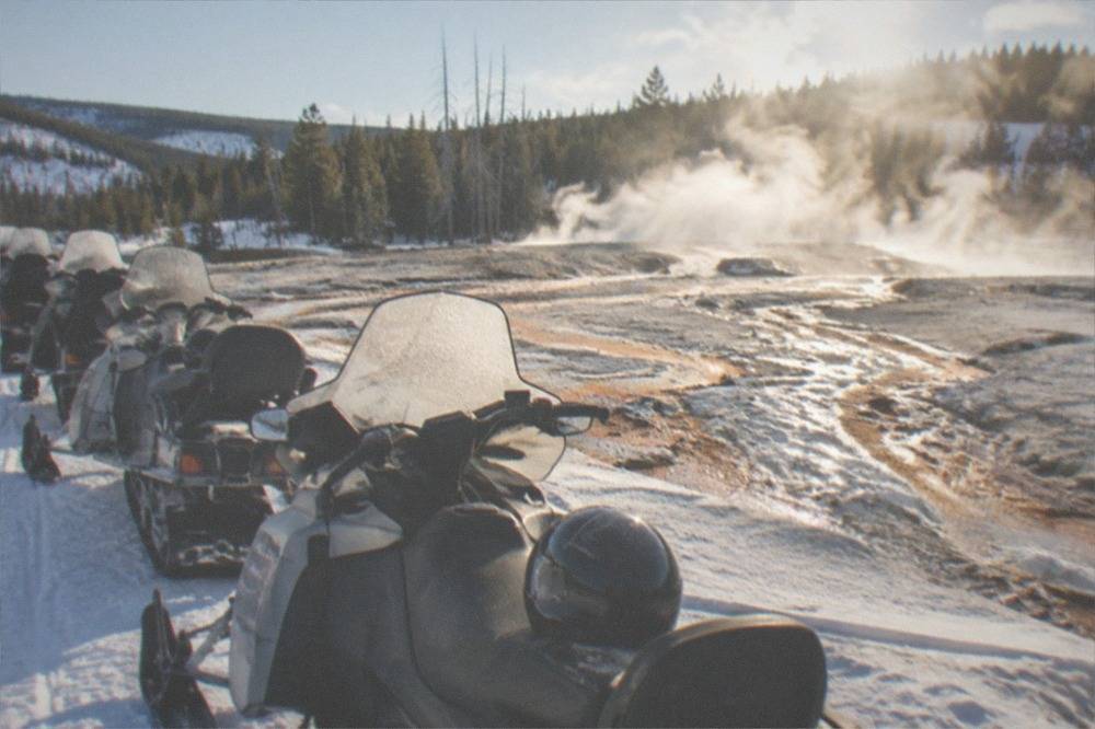 Line of parked snowmobiles on a groomed snowy road beside steaming thermal pools in early morning light