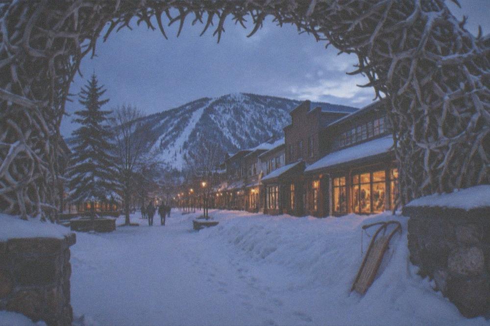 Small western town main street at dusk with snow banks along the storefronts and warm window lights.