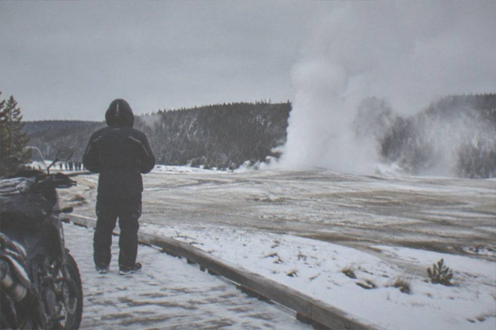 A snowmobile rider in an insulated suit waiting beside a geyser boardwalk under gray skies