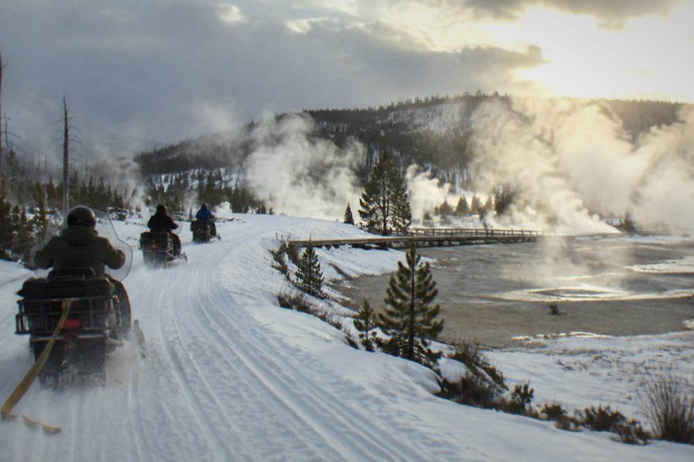 Snowmobile group riding a groomed road past steaming thermal pools in winter