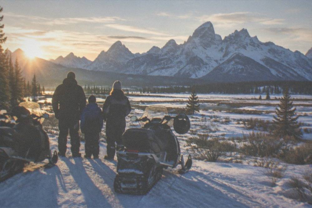 Family in snowsuits and helmets standing beside parked snowmobiles on a sunny winter trail