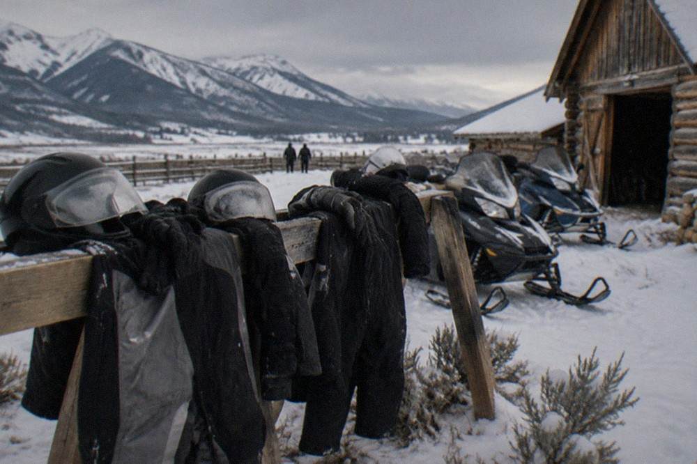 A row of parked snowmobiles with helmets and insulated suits laid out on a wooden rack beside a snow-covered ranch building