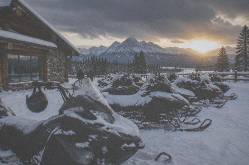 A parking lot of snowmobiles for rent lined up outside a rental shop under deep snow and mountain peaks