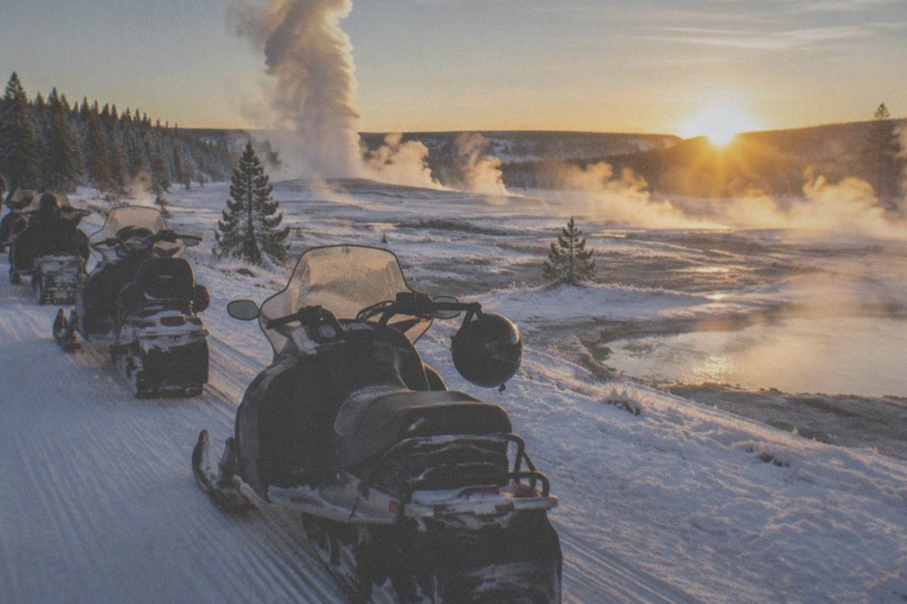Snowmobiles parked on a groomed white road beside steaming geothermal pools in a snowy valley