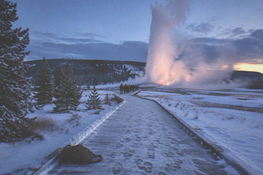 A tall geyser erupting steam into cold air above a snow-covered basin ringed by boardwalks