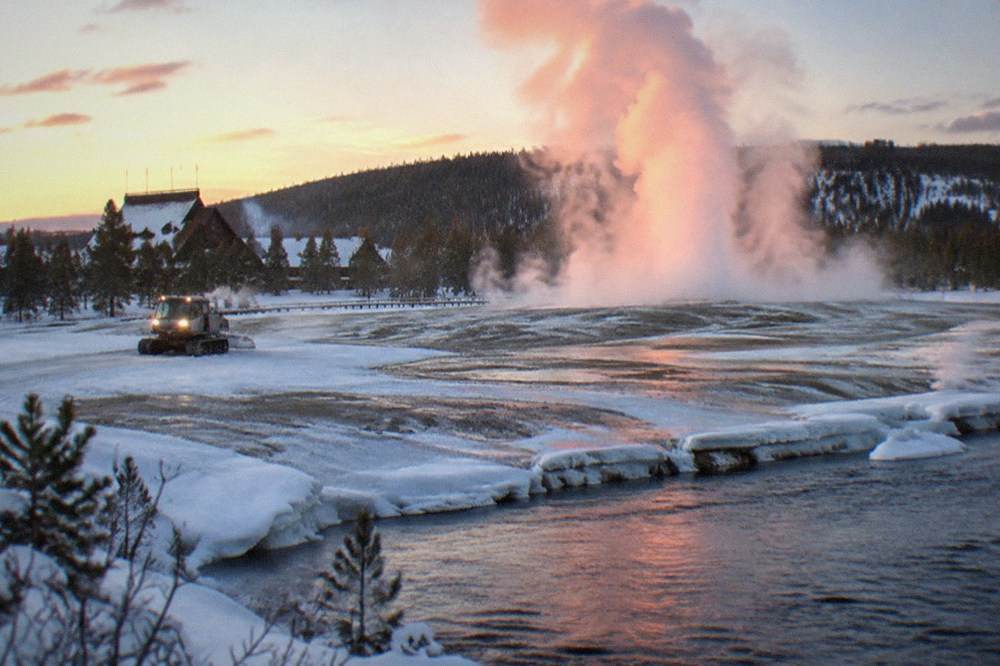 Geyser steam rising from a snow-covered thermal basin beside a groomed winter road