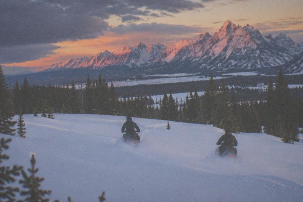 Two snowmobiles carving through open powder in a mountain meadow under tall peaks