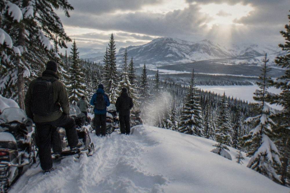 A guided snowmobile group paused on a snowy forest road with deep drifts along the edge