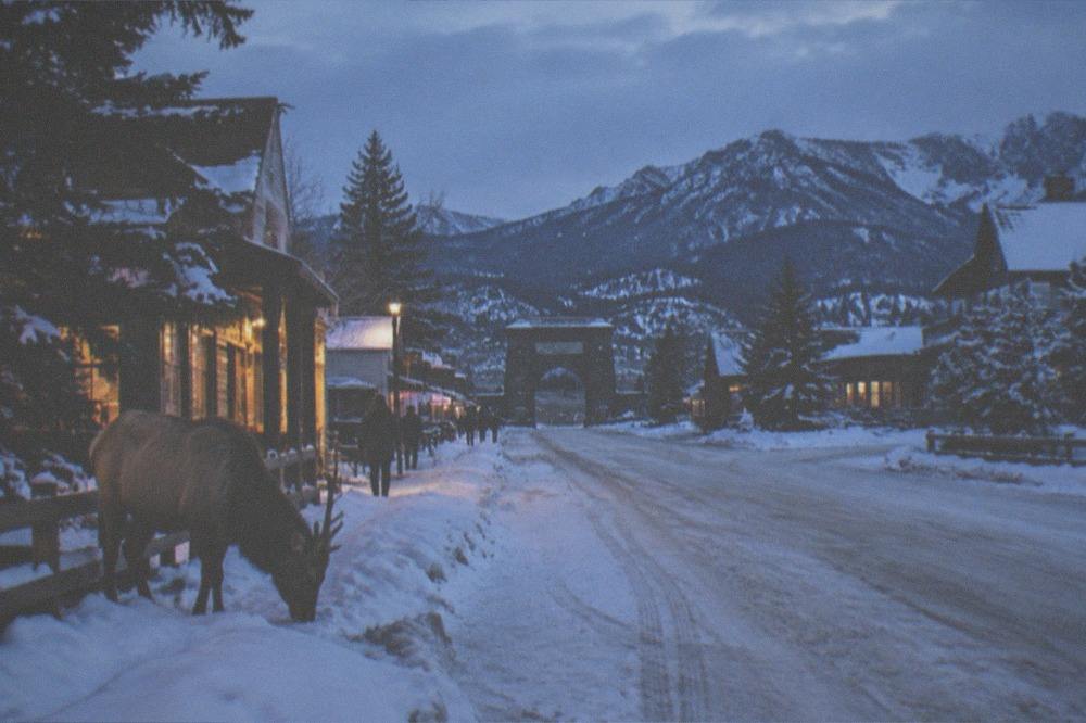 A snowy small-town main street at dusk with warm lights glowing in shop windows and mountains behind