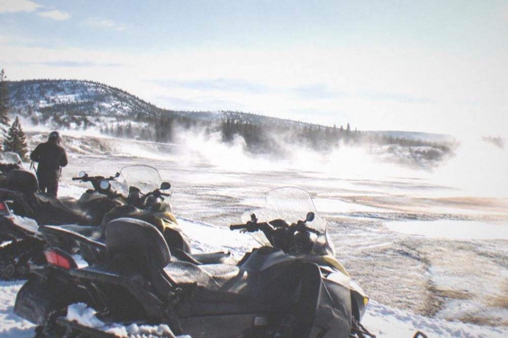 Group of snowmobiles parked in a row beside steaming geothermal basin in snow