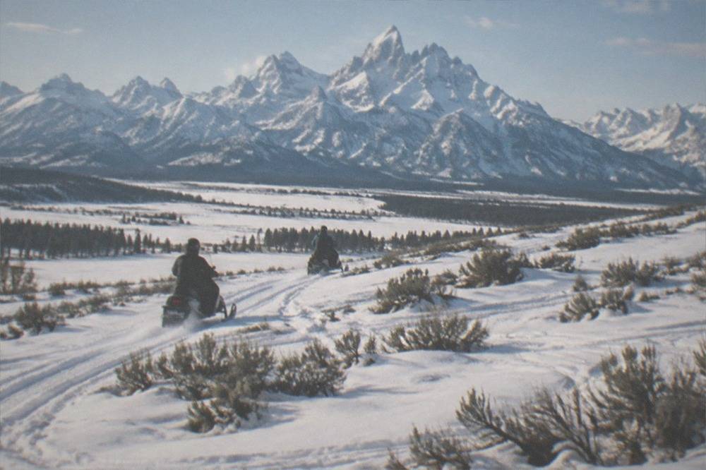 Two snowmobiles climbing a powder-covered meadow slope beneath jagged snow-capped mountain peaks